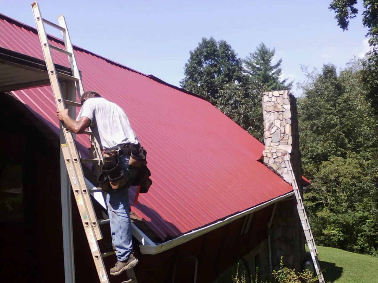 Roofer on a ladder beside a red metal roof and stone chimney — Soffit & Fascia Repair by Roofing Brothers in Lynwood